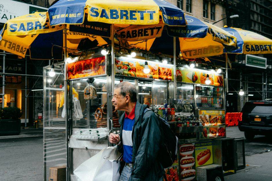 Elderly man passing a New York City food stall featuring Sabrett hot dogs on a lively city street.